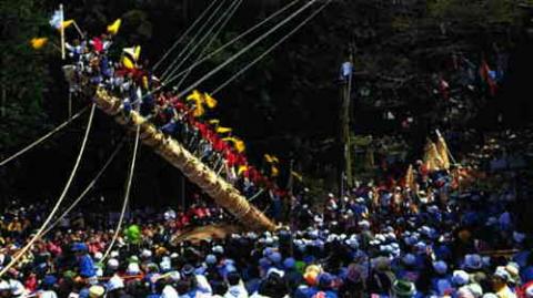 複数の綱が張り巡らされた巨大な柱が地面に斜めに傾いており、その上に多くの人が集まって旗を振る様子を群衆が取り囲んでいる御柱祭の写真