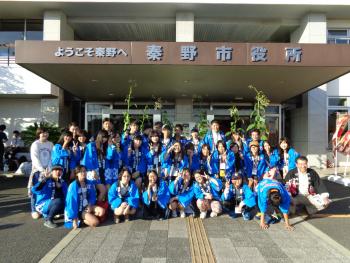 「秦野市役所」の玄関前で、青い法被のような上着を着た30人ほどの若者たちが、楽しそうに笑顔でポーズをとっている集合写真