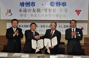 「坡州（パジュ）市・秦野（はだの）市 永遠の友情/영원한 우정」と書かれた横断幕の下でスーツ姿の4人の男性がテーブルの前に立っており、中央の2人が協定書を持って握手し、両脇の2人は拍手をしている調印式の写真