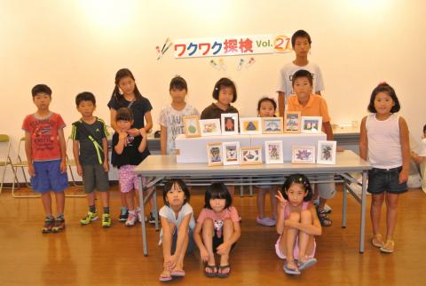 「ワクワク探検 Vol.27」と書かれた横断幕の前で、10人以上の子供たちが制作したと思われる小さな作品を展示したテーブルを囲み、記念撮影している写真