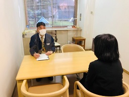 白いマスクをつけた相談員の男性が、向かいに座る女性と向き合い、手元の書類に何かを書き込んでいる様子を撮影した相談室内の写真