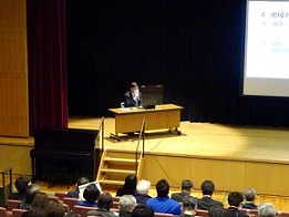 資料が映し出されたスクリーンの舞台の左側のモニターが設置された机に座った男性が話しているのを、聴衆が熱心に話を聞いている講演会の様子の写真