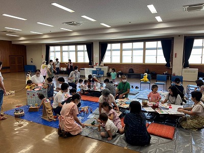 施設の広い洋室に敷かれた青いビニールシートの上で、親子連れが大勢集まり、座卓を囲んで絵を書いたり、おもちゃや工作、絵本などで遊んだり、交流したりしている様子の写真