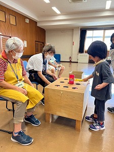 公民館のような広間で、黄色のエプロンをつけた高齢の女性と別のスタッフが見守る中、小さな男の子が木製の叩くおもちゃで遊んでいる、地域の子育て支援の一コマを捉えた写真