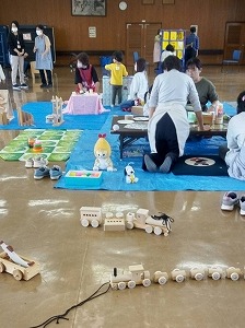 紐で連結された木製の電車のおもちゃが床に置かれた室内で、青いビニールシートの上で複数の親子やスタッフが遊具を囲んで交流する様子を捉えた写真