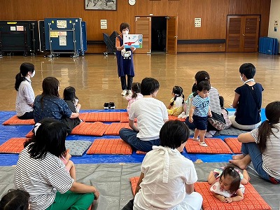 広いホールの床に敷かれた青いシートとオレンジ色の座布団の上で、小さな子供連れの母親たちが、中央に立つエプロン姿の女性が絵本を広げて読み聞かせをしているのを熱心に聞いている写真