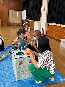 ホールの床に敷かれた青いビニールシートの上で、2人の幼い女の子が、段ボールで作られたおままごと用のキッチンテーブルを囲み、座り込んでサポートする大人たちと一緒に、笑顔でおもちゃの野菜や果物を使って遊んでいる写真