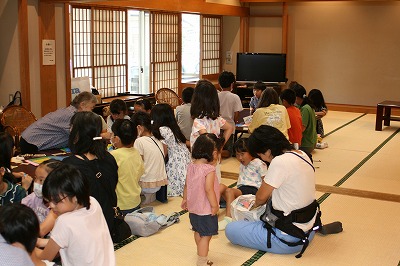 奥には障子や襖、テレビが設置された和室の折り紙コーナーで、大勢の子供たちと数名の保護者やスタッフが座卓を囲んで座り、折り紙を楽しんだりしている、地域交流イベントの風景写真