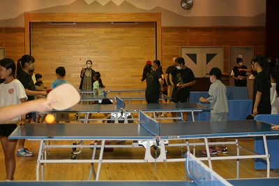 複数の卓球台が設置された体育館のような広い木目の室内の卓球コーナーで、子供から大人まで大勢の参加者がラケットを手に卓球を楽しんでいる様子を捉えた写真