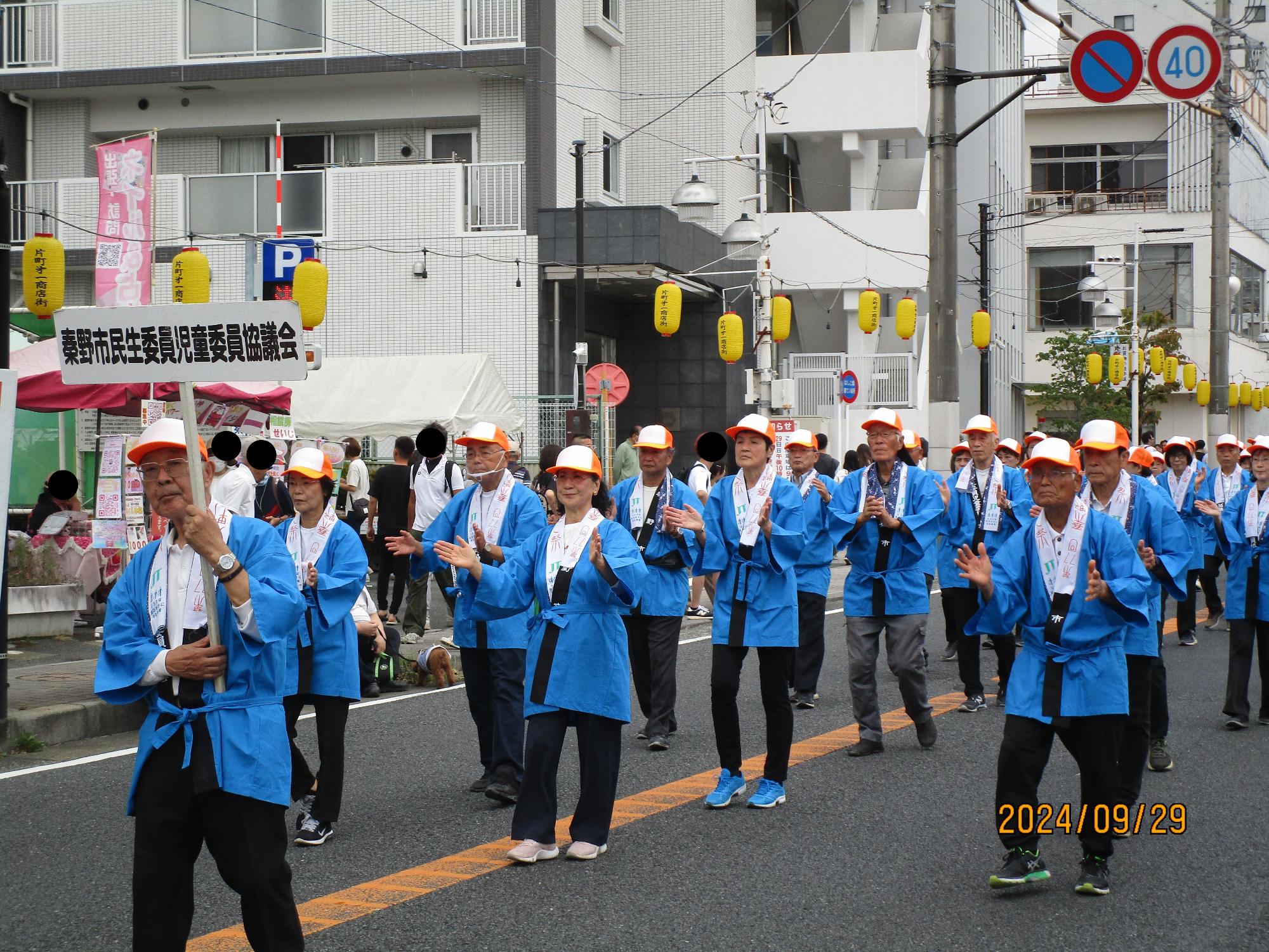 青い法被とオレンジ色の帽子をかぶった大勢の人々が、路上に吊るされた提灯の下で、拍手をしながら行進している写真