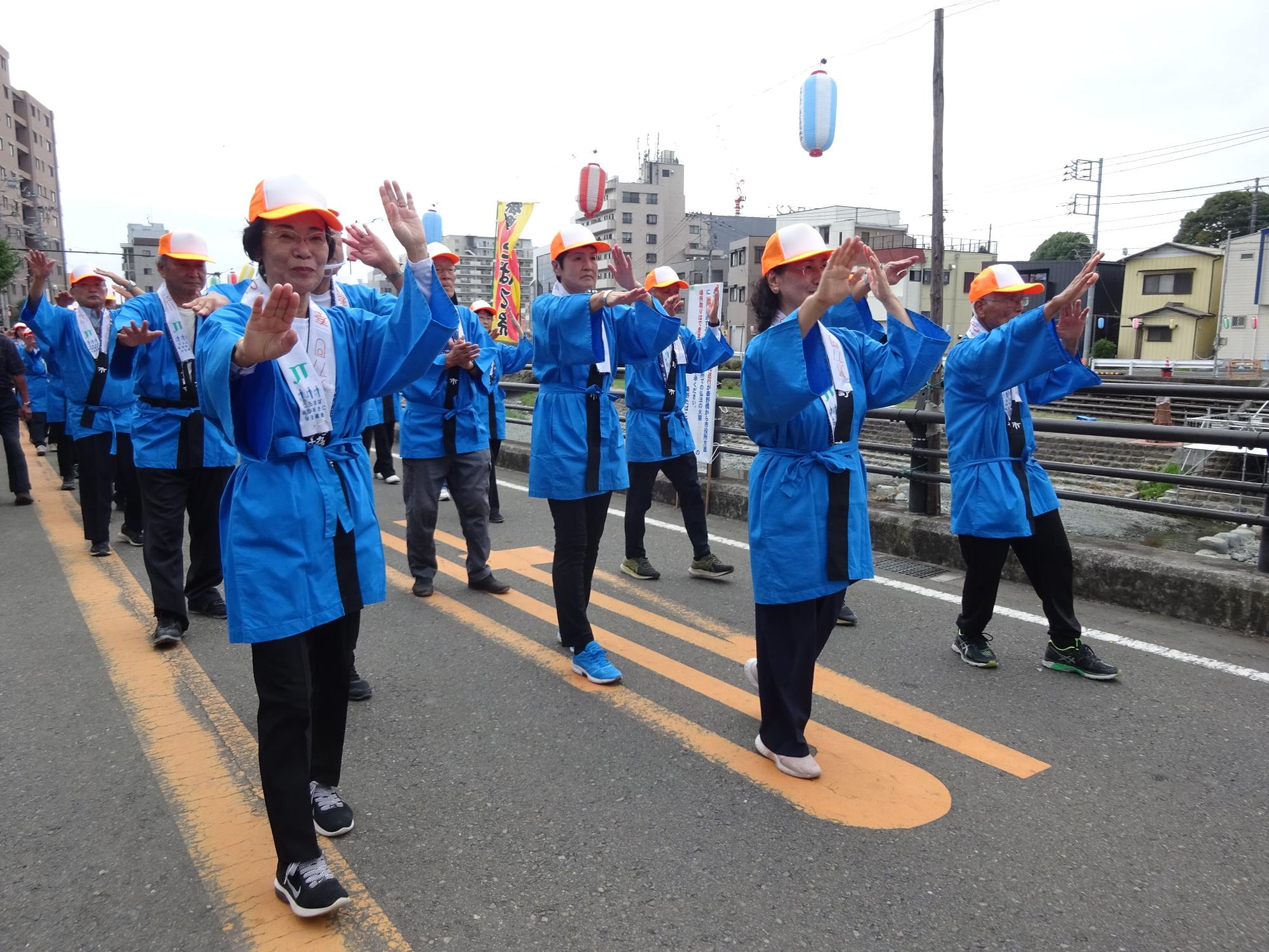 青い法被とオレンジ色の帽子を身につけた大勢の人々が、路上で両手を挙げながら踊りながら行進している祭りの写真