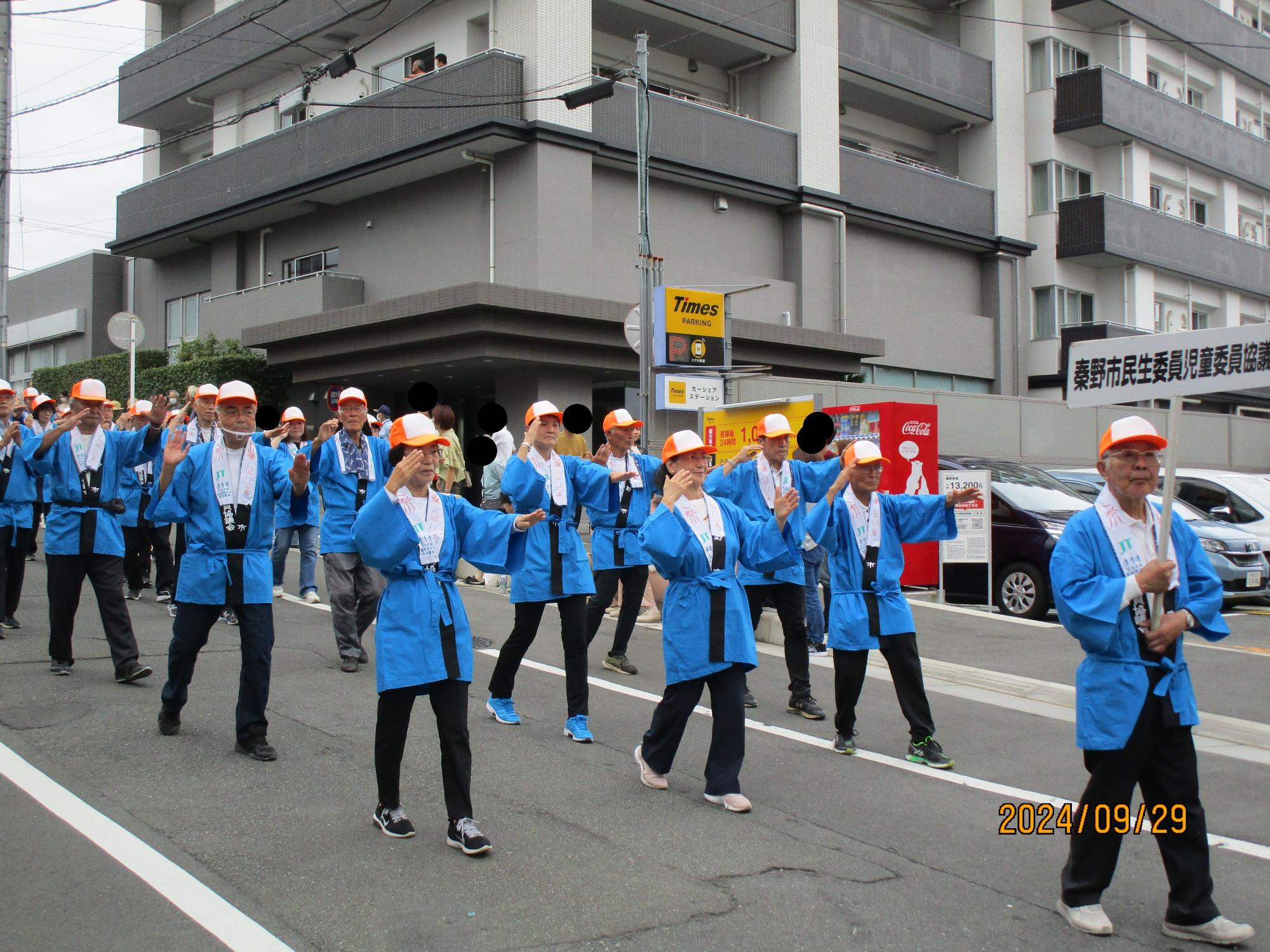 祭りのパレードで、青い法被とオレンジ色の帽子を着用した大勢の人々が、笑顔で手を振りながら道路を行進している写真