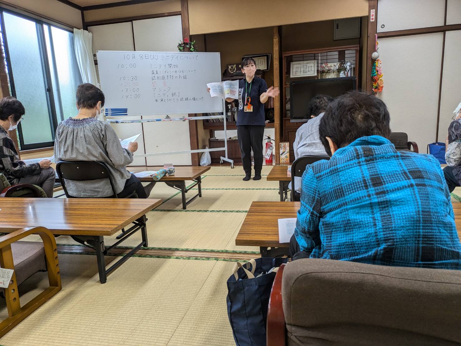 和室に敷かれた畳の上で、数名の高齢者が低い座卓に向かい合って座り、中央で若い女性の担当者がホワイトボードの前で身振り手振りで話しをしている様子の写真
