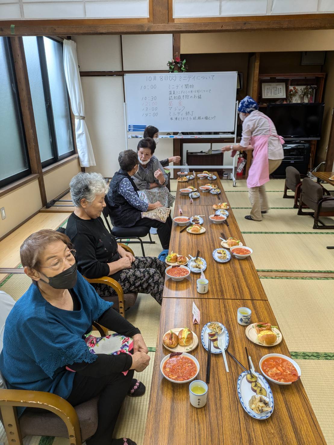 奥のホワイトボードには今後の予定が書かれた和室で、トマトソースのパスタのような料理や2種類の具が入ったロールパンが置かれたテーブルを囲んで座った数名の高齢者が談笑しており、、ピンクのエプロンをつけたスタッフが料理を配膳している様子の写真