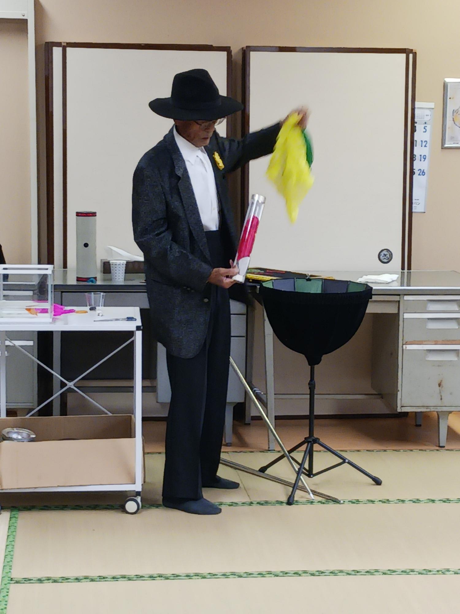 和室に敷かれた畳の上で、黒いハットとスーツを着用した高齢の男性が、小道具が置かれた机と三脚付きの黒いボウルを前に、黄色いハンカチを使ってマジックを披露している様子の写真