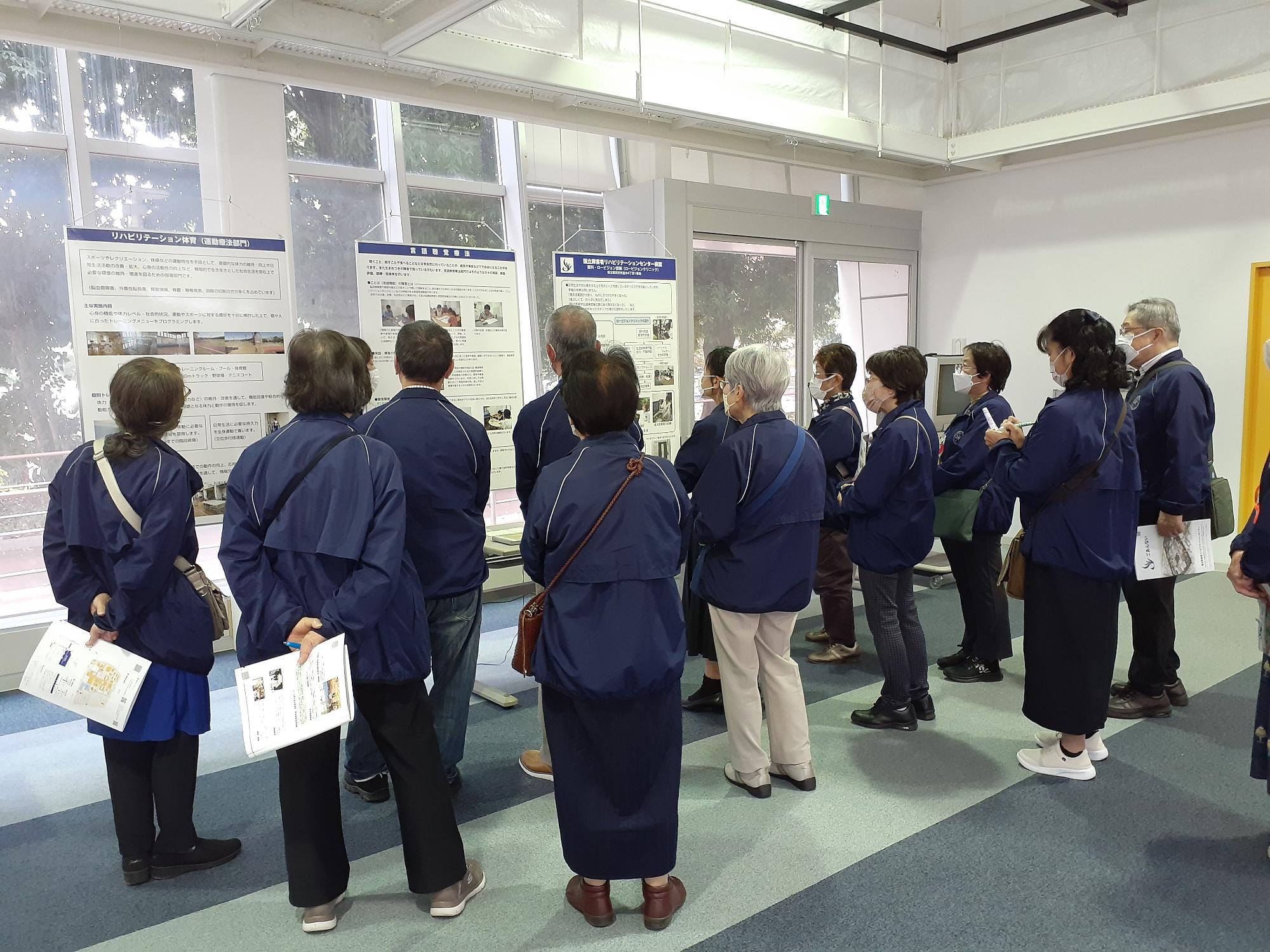 濃紺の上着を着た大勢の人々が、部屋に展示された複数のパネルを熱心に鑑賞している視察研修の写真