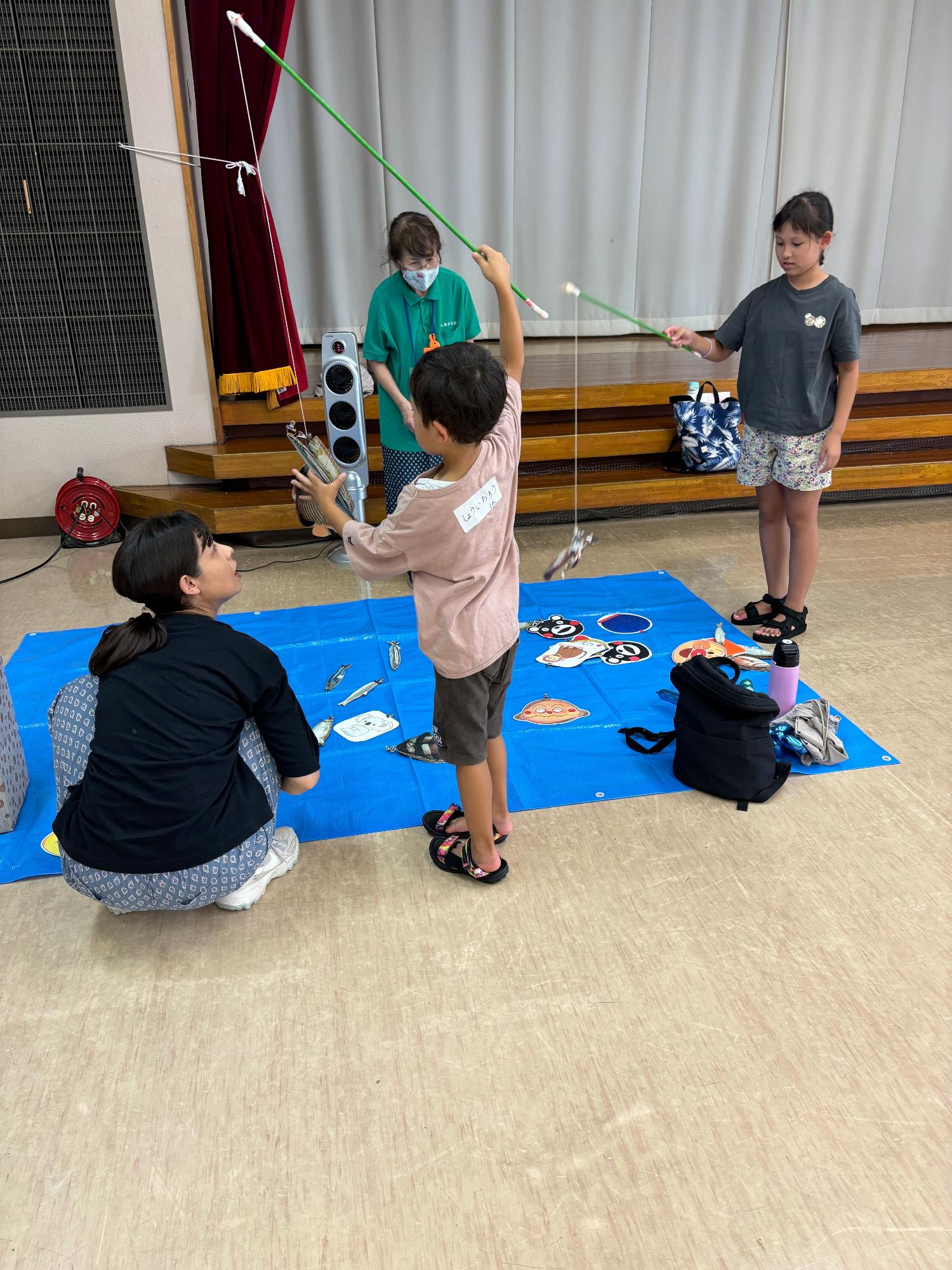 屋内のステージ前で、青いシートの上に置かれた魚の絵を釣り竿で釣るゲームを、数人の子供たちが大人に見守られながら楽しんでいる写真