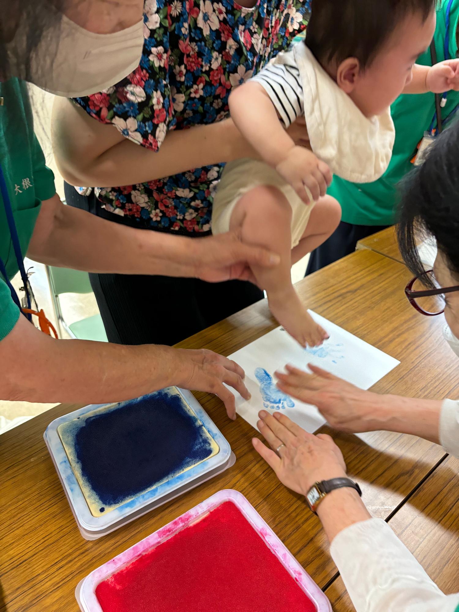 大人たちが協力して赤ちゃんを支えながら、青いインクをつけた足型を白い紙にスタンプしているイベントの様子を捉えた写真