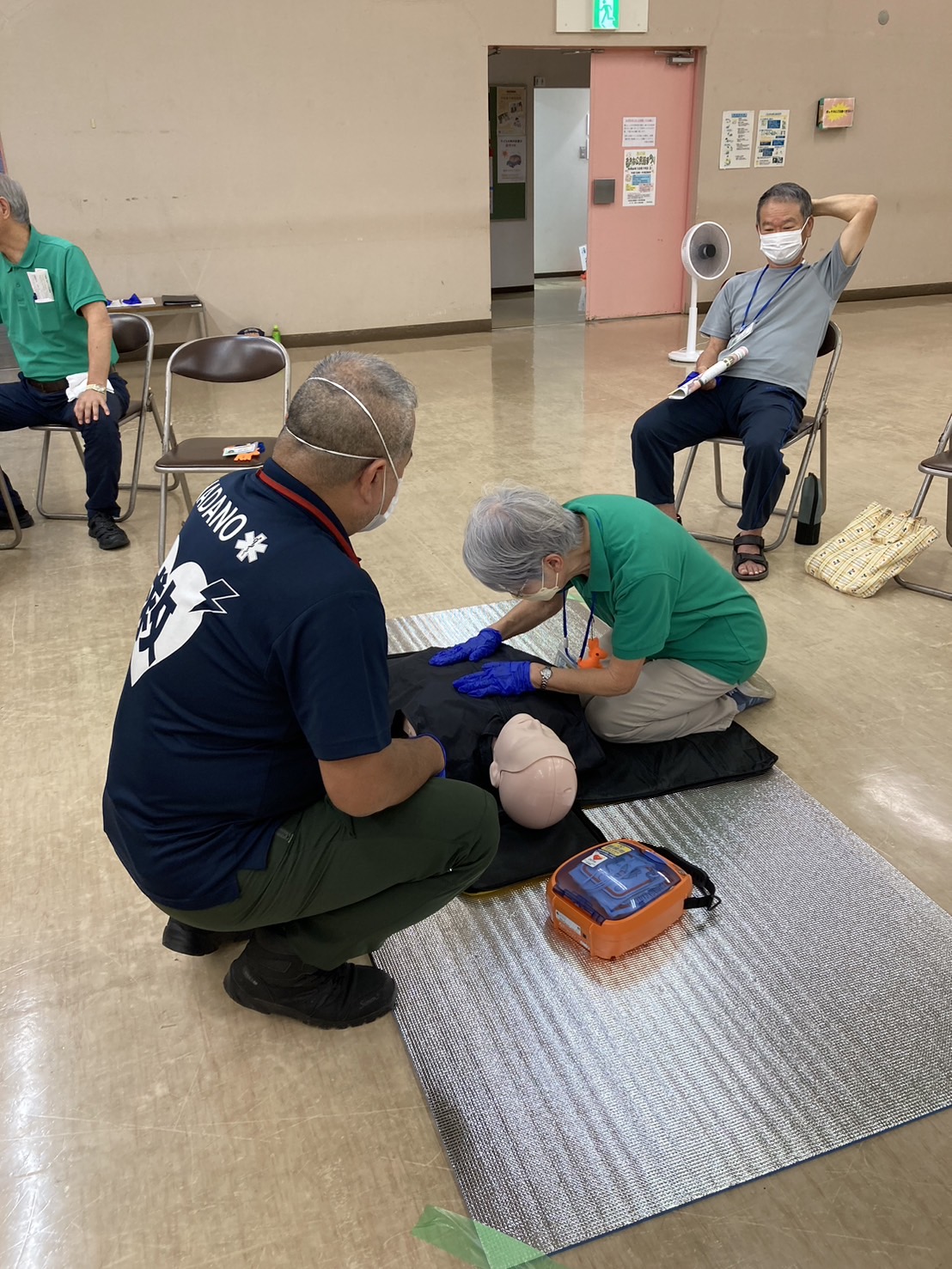 救急救命講習会に参加した女性が、心肺蘇生訓練用の人形を使い、指導役の男性からアドバイスを受けながら胸骨圧迫を行っている様子の写真