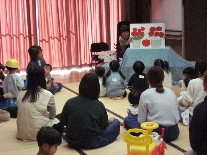 畳の部屋で、子どもたちが大人に囲まれて座り、絵本の読み聞かせを静かに聞いている様子を写した写真