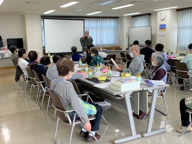 広間に集まったマスク姿の参加者たちが、中央に立つハーモニカ演奏を熱心に聞いている様子の写真