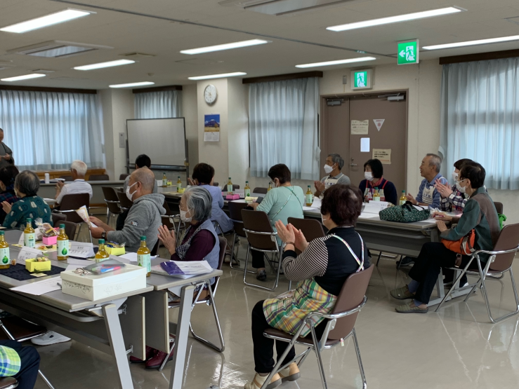 広い会議室でマスク姿の高齢者たちが会議机の周りに座り、中央で誰かの話に耳を傾けながら拍手をしているゲームの様子の写真