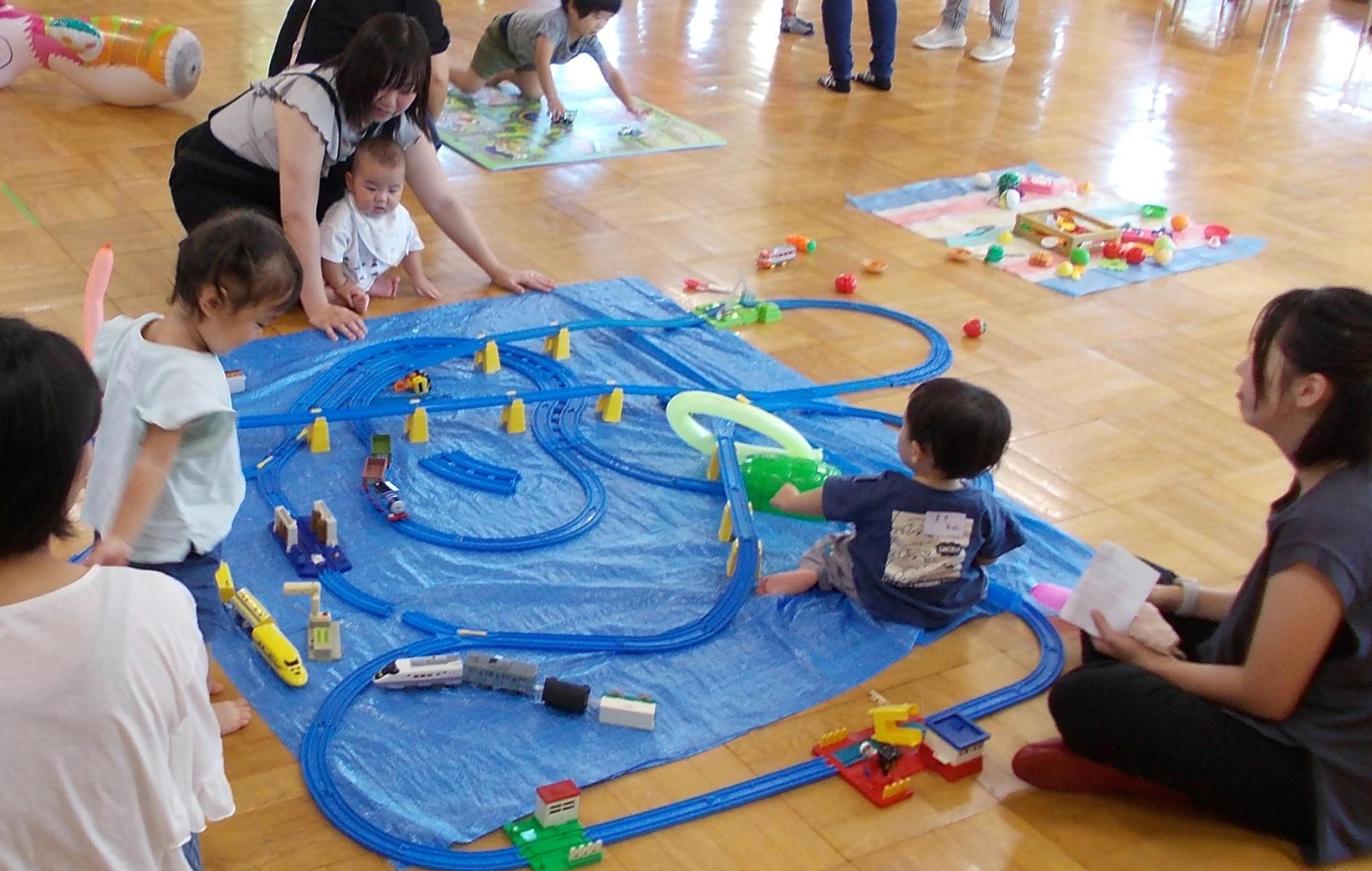 木の床の広い室内で、青いビニールシートの上で幼い子供たちが、母親や他の大人が見守る中、プラスチック製の青いレールを組み合わせて遊ぶ電車のおもちゃで夢中になって遊んでいる様子の写真