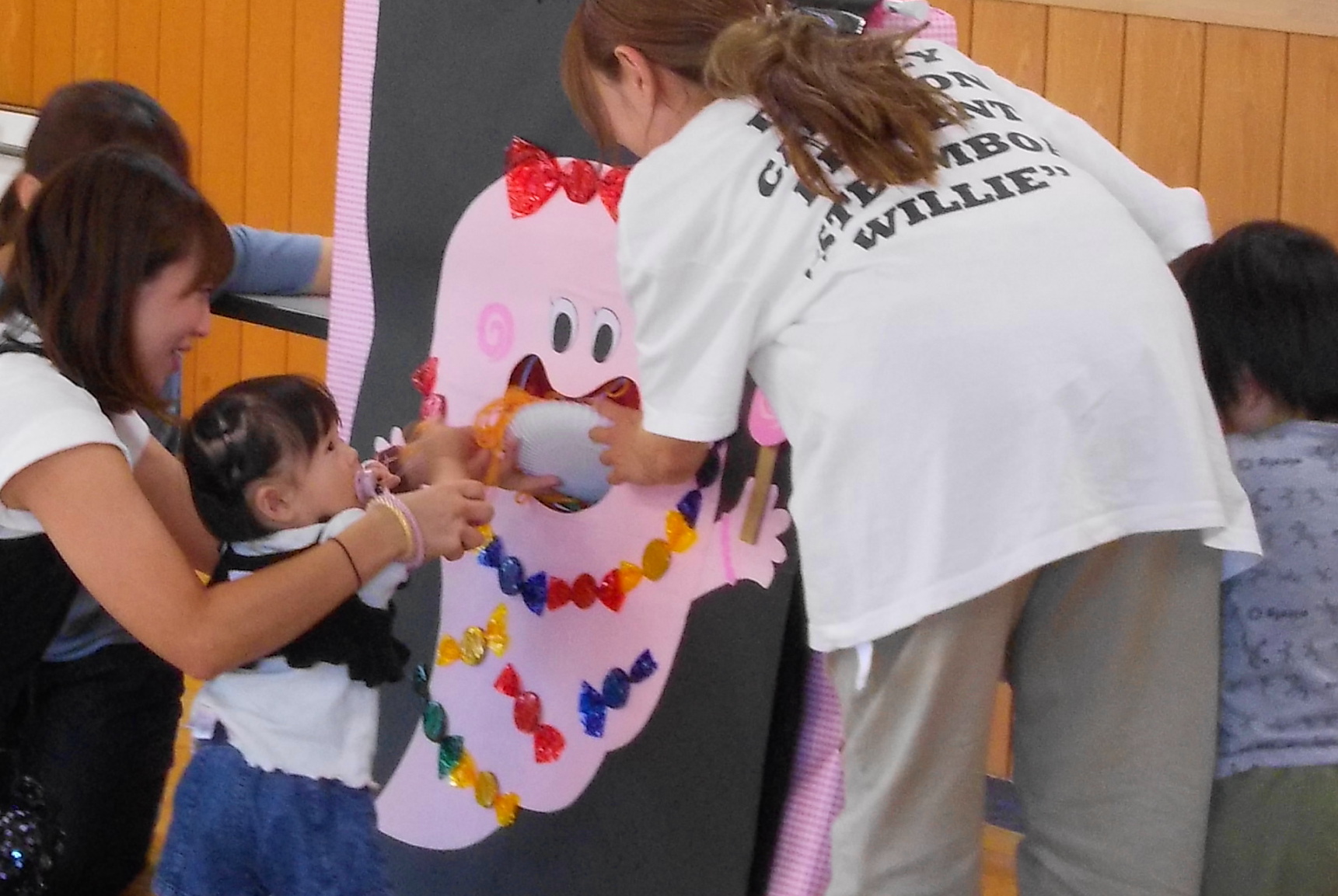 大人の女性たちに見守られながら、可愛らしいピンク色のオバケのイラストが描かれたパネルに、幼い女の子が手を入れて楽しんでいる様子の写真