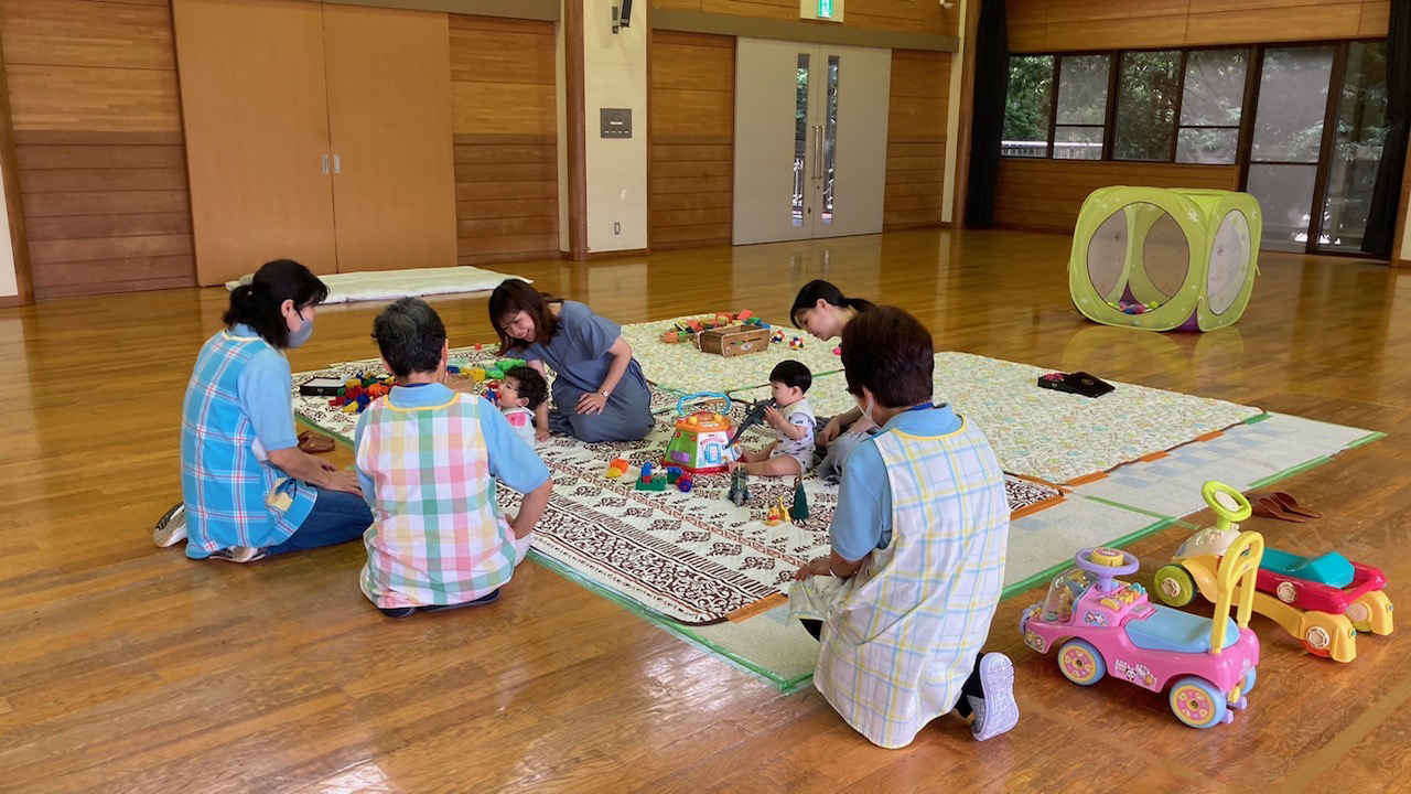 木目の床が広がる室内で、数人の大人と子供たちが、ラグマットの上におもちゃを広げ、交流会に参加している風景を捉えた写真