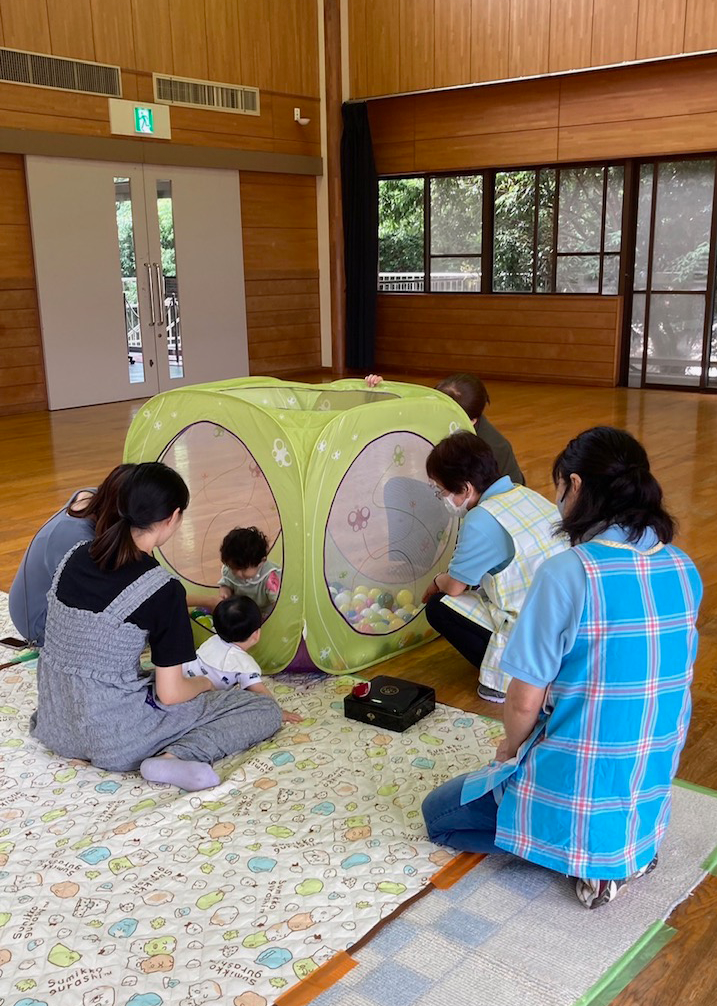 床の上に敷かれたシートの上で、複数の大人と子供たちが、ボールプールが設置された緑色のプレイテントを囲んで楽しんでいる子育てイベントの様子の写真