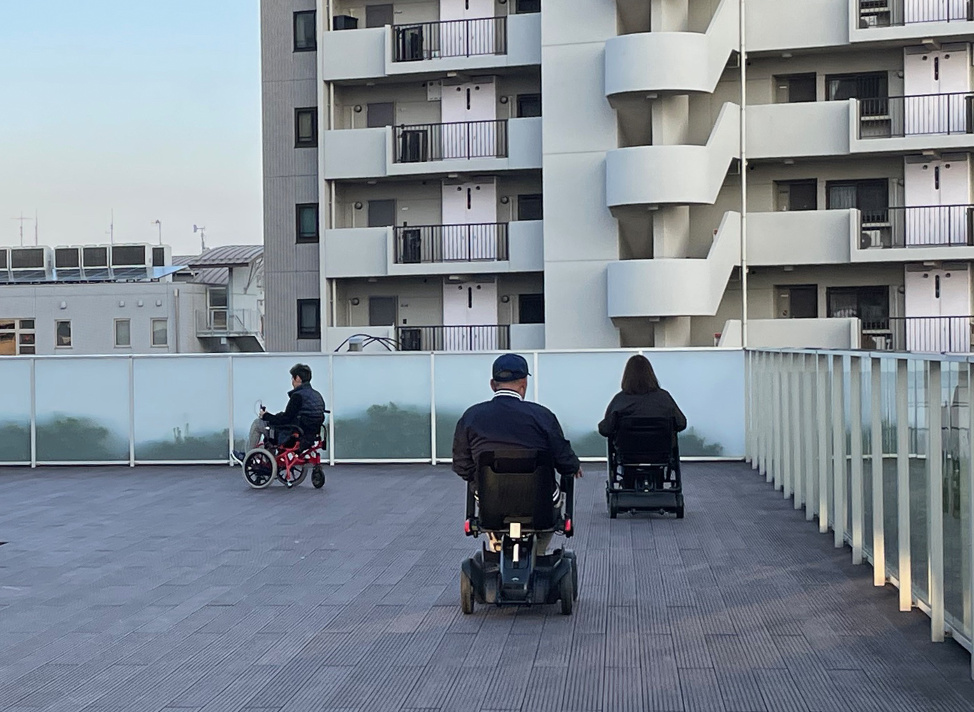 周囲を集合住宅の建物に囲まれた、建物の屋上のような広い場所で、電動車いすに乗った3人が試乗を行っている様子を写した写真