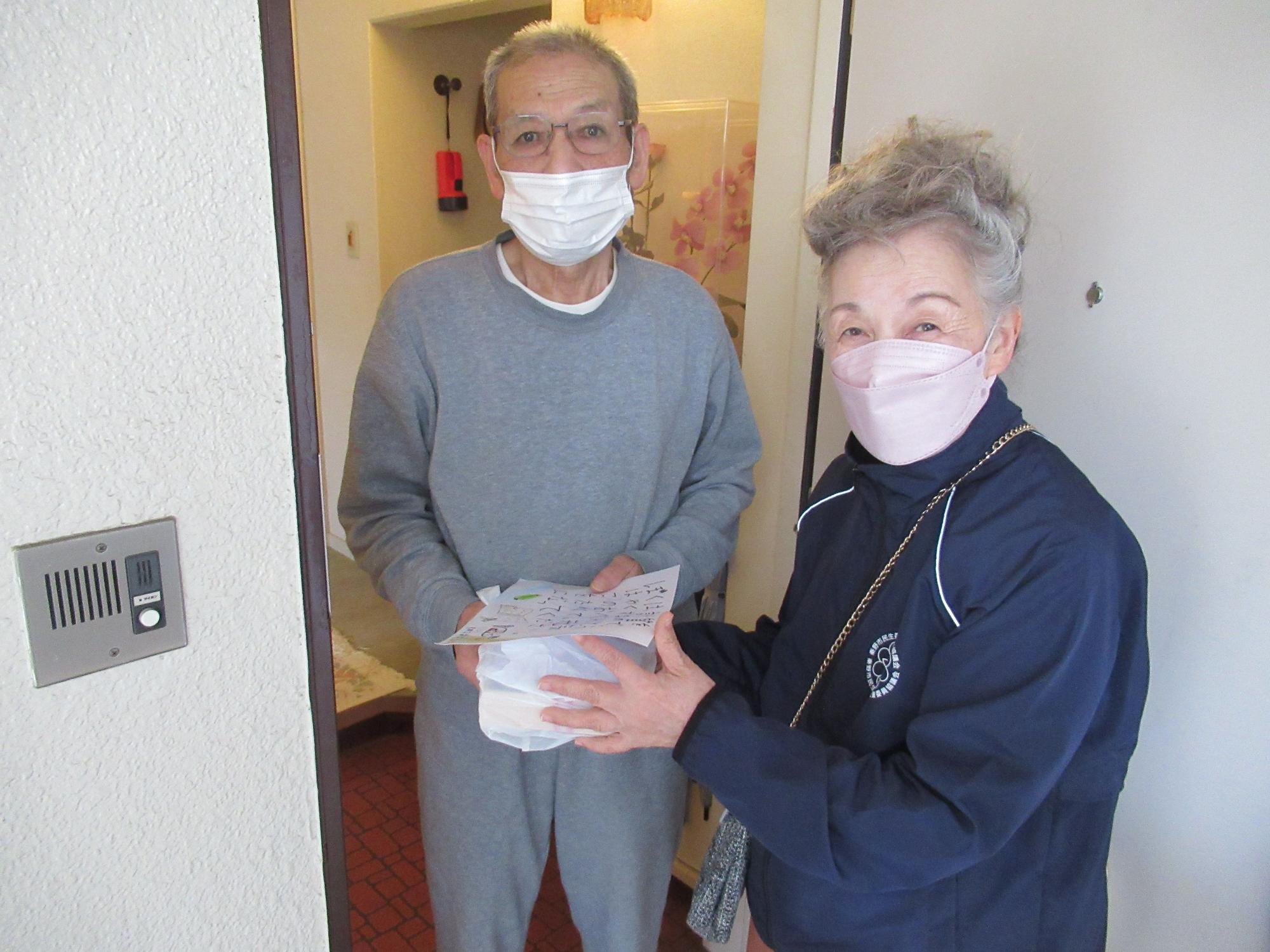 インターホンが設置された玄関で、マスクをつけた高齢の女性が、手書きのメッセージが添えられた弁当を、住人の男性に手渡している地域交流の様子を捉えた写真