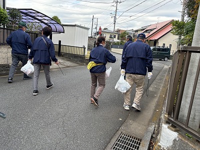 複数の中高年が紺色のジャンパーを着て、ゴミ袋と火ばさみを持って道路を歩き、清掃活動を行なっている、地域美化活動を捉えた写真