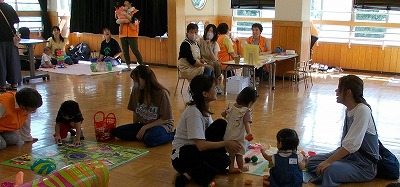 公民館のような広いフローリングの室内で、大人の女性たちが数人の乳幼児たちと一緒に床に座り、おもちゃで遊んで交流しており、奥のテーブルでは他のスタッフらしき人々が座っている、子育て支援の風景写真