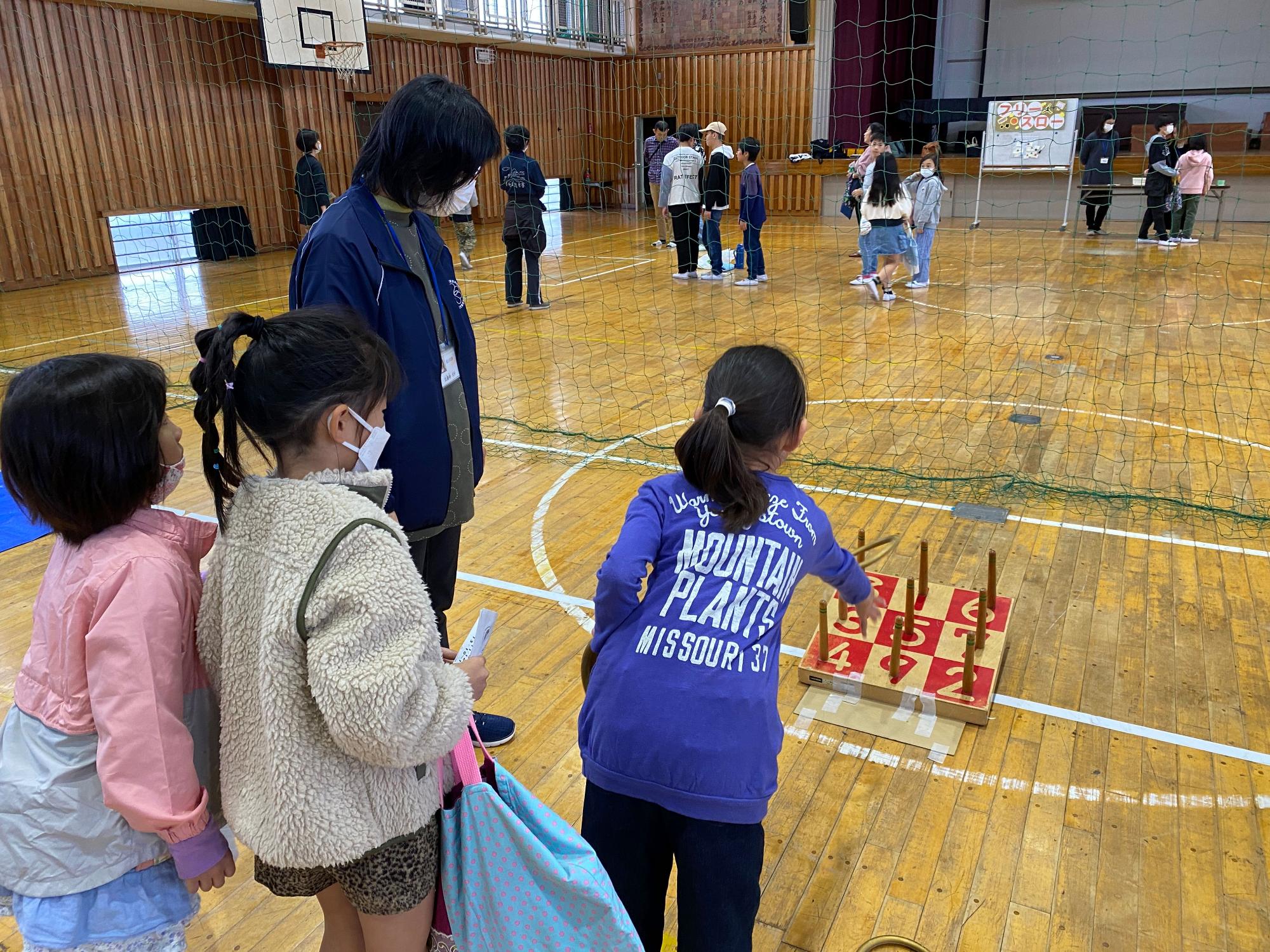 体育館で3人の女の子たちが木の台にある棒を数字の書かれた穴に輪っかを投げて遊んでおり、その様子を大人が見守っている写真