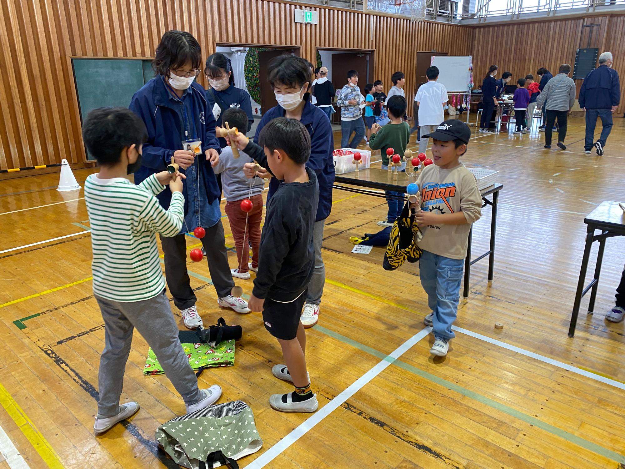 体育館で子供たちがけん玉をして遊んでおり、大人が子供たちにやり方を教えている様子を撮影した写真