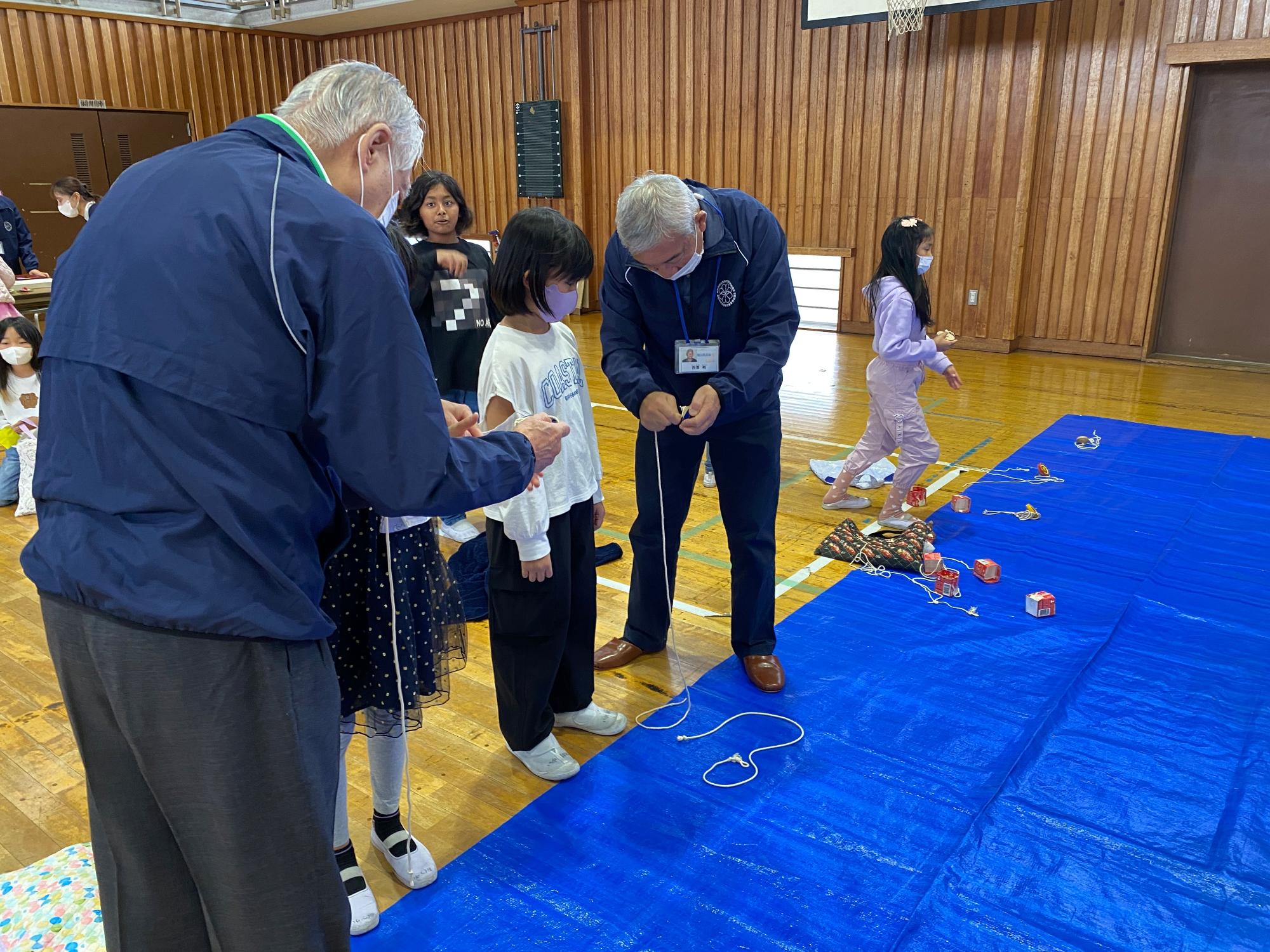 体育館の床に敷かれた青いシートの上で、大人たちが子供たちにコマに糸を巻くやり方を教えながら、一緒に遊んでいる様子を撮影した写真
