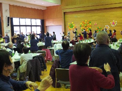 小さなステージがある室内で、紺色のジャンパーを着たスタッフやマスクを着用した大勢の高齢者がテーブルの周りに立ち、両手を挙げて踊っている様子の写真