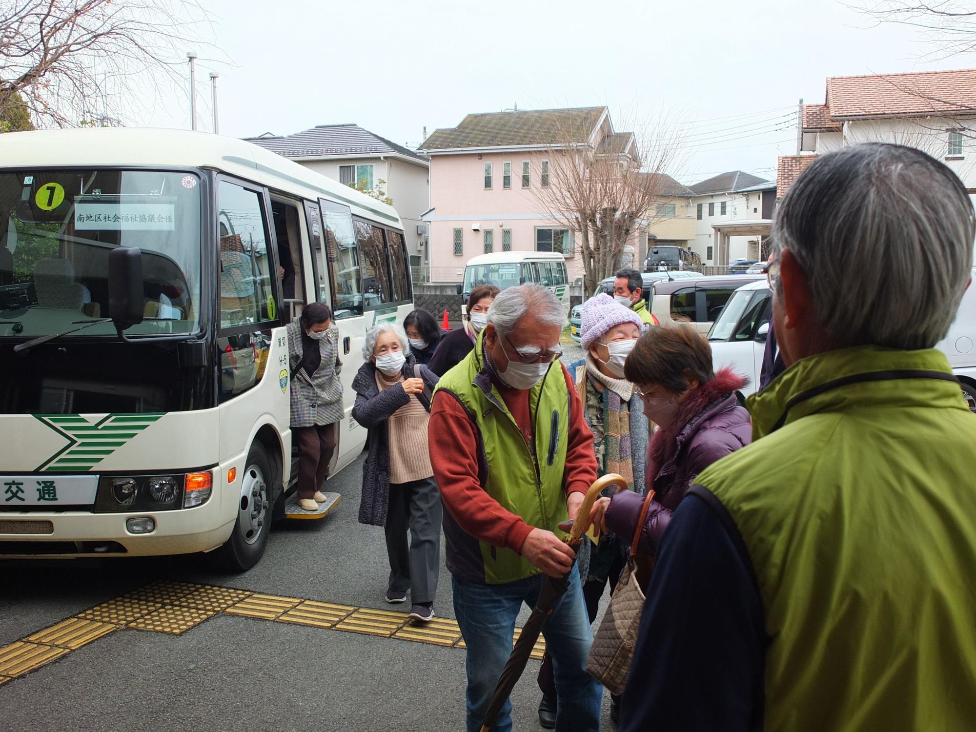 傘や杖を持った大勢のマスクを着用した高齢者が、中型の送迎バスから降りてきている様子の写真