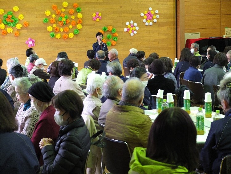 木目の壁にはカラフルな花の装飾が施されている屋内で、お茶のペットボトルが置かれたテーブルの席に座ったマスクを着用した大勢の参加者が、正面で挨拶している一人の女性の話を熱心に聞いている様子の写真