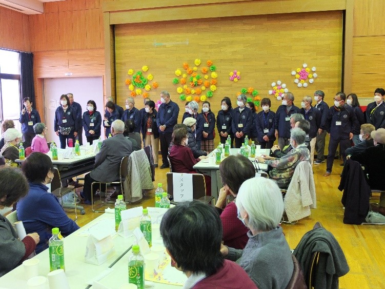 集会所の木目調の壁を背景に、紺色のスタッフジャンパーを着てマスクを着用したスタッフが横一列に並び、手前のテーブルでは大勢の高齢者が緑茶のペットボトルを前に座って、左端に立っている女性の話を傾聴している様子の写真