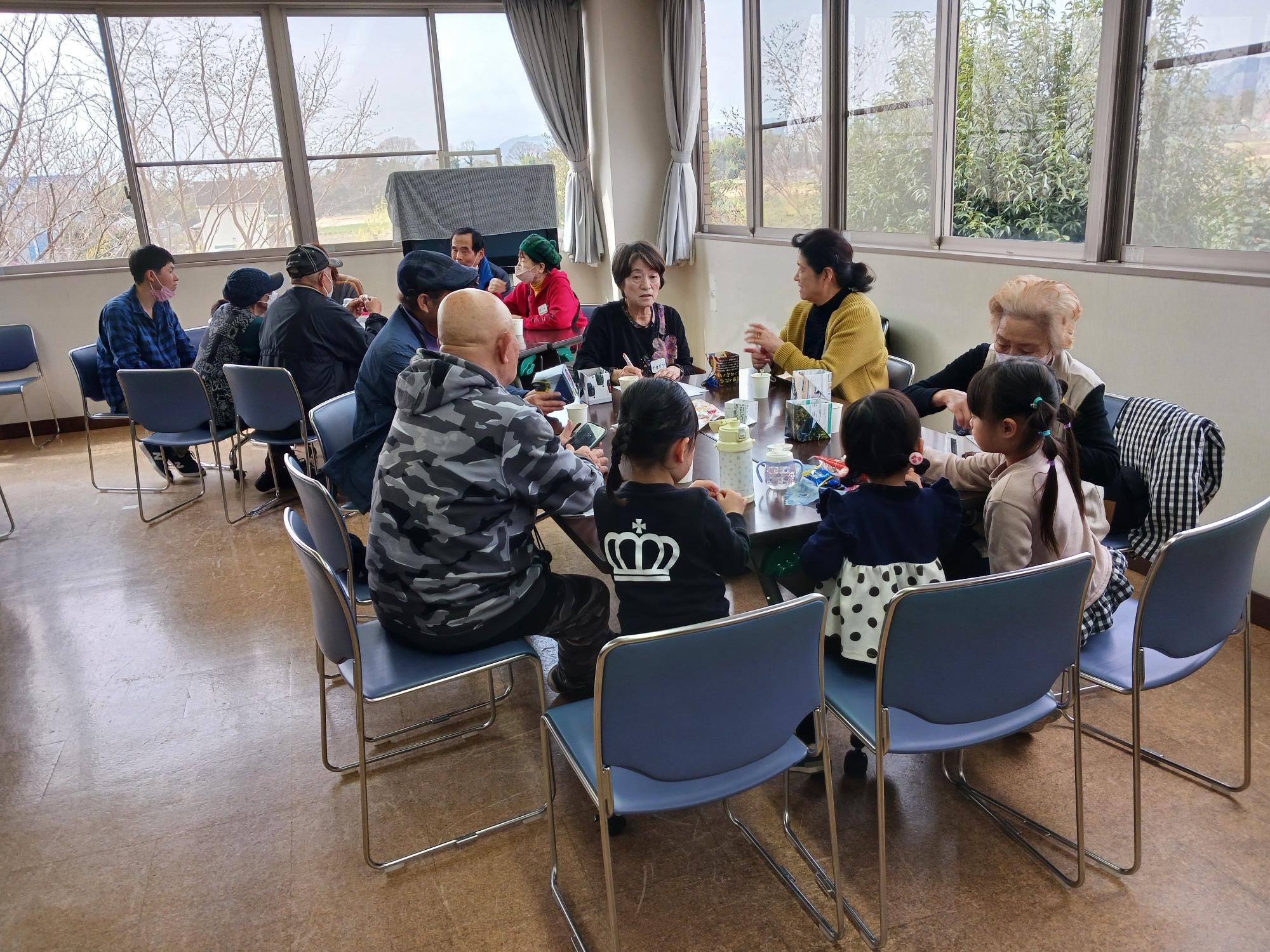 採光の良い広間で、高齢者と中年の大人、そして数人の小さな子供たちが二つのテーブルに分かれて座り、お茶を飲んだり会話をしたりしている、世代間交流の場を映した写真