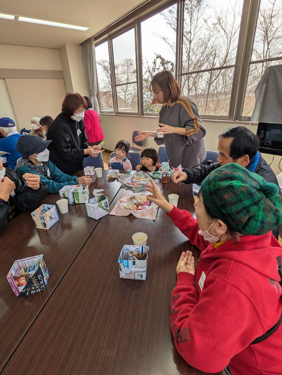 広くて明るい室内で、一組のテーブルを囲んでマスクを着用した高齢者や大人たち、2人の幼い子供連れの親子が座り話をしている、世代を超えた地域交流会の写真