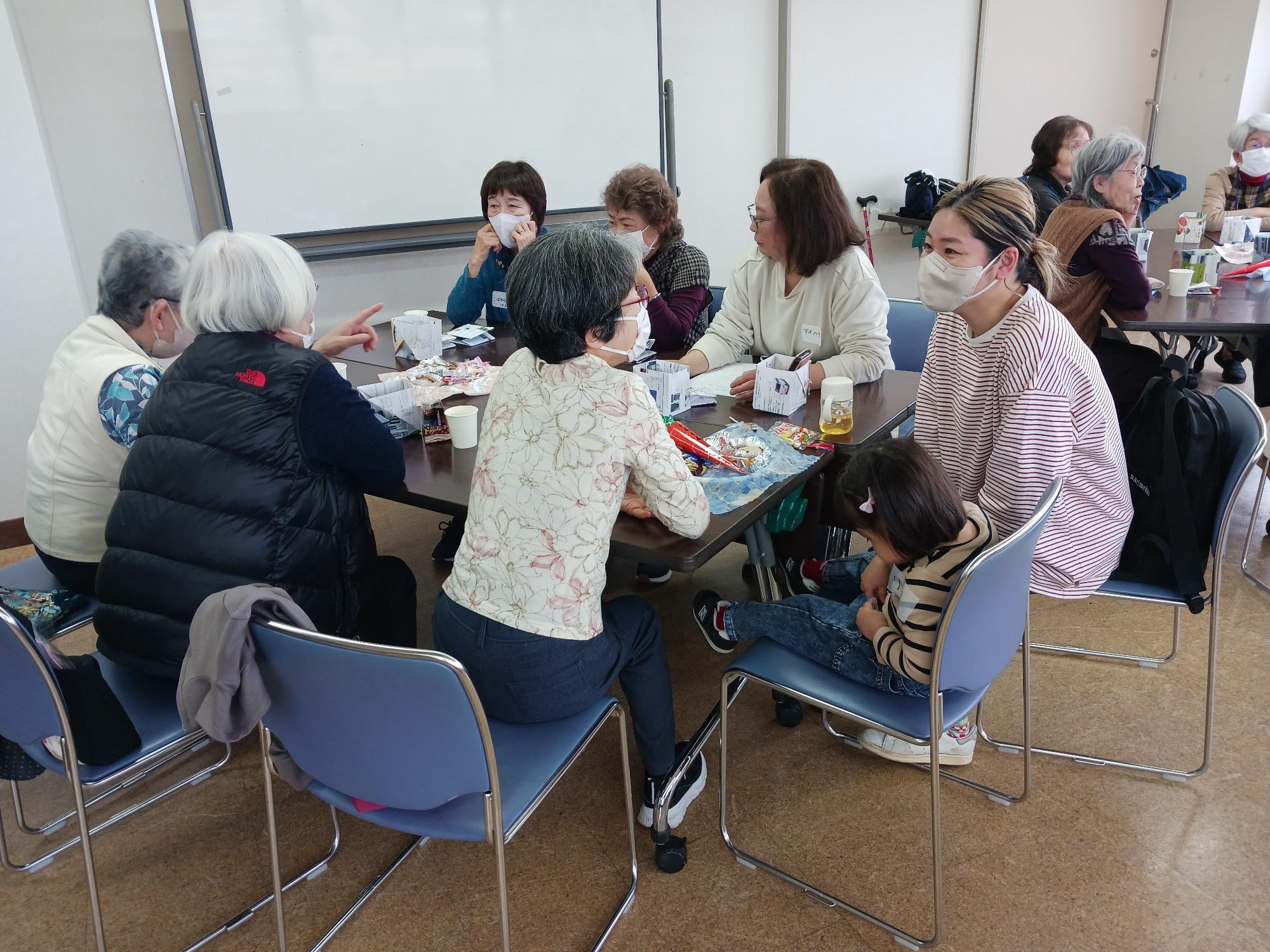 公共の集会室のような場所で、複数の高齢の女性と子供連れの若い女性が、飲み物やお菓子が並んだテーブルを囲んで会話を楽しんだりしている地域交流の様子の写真