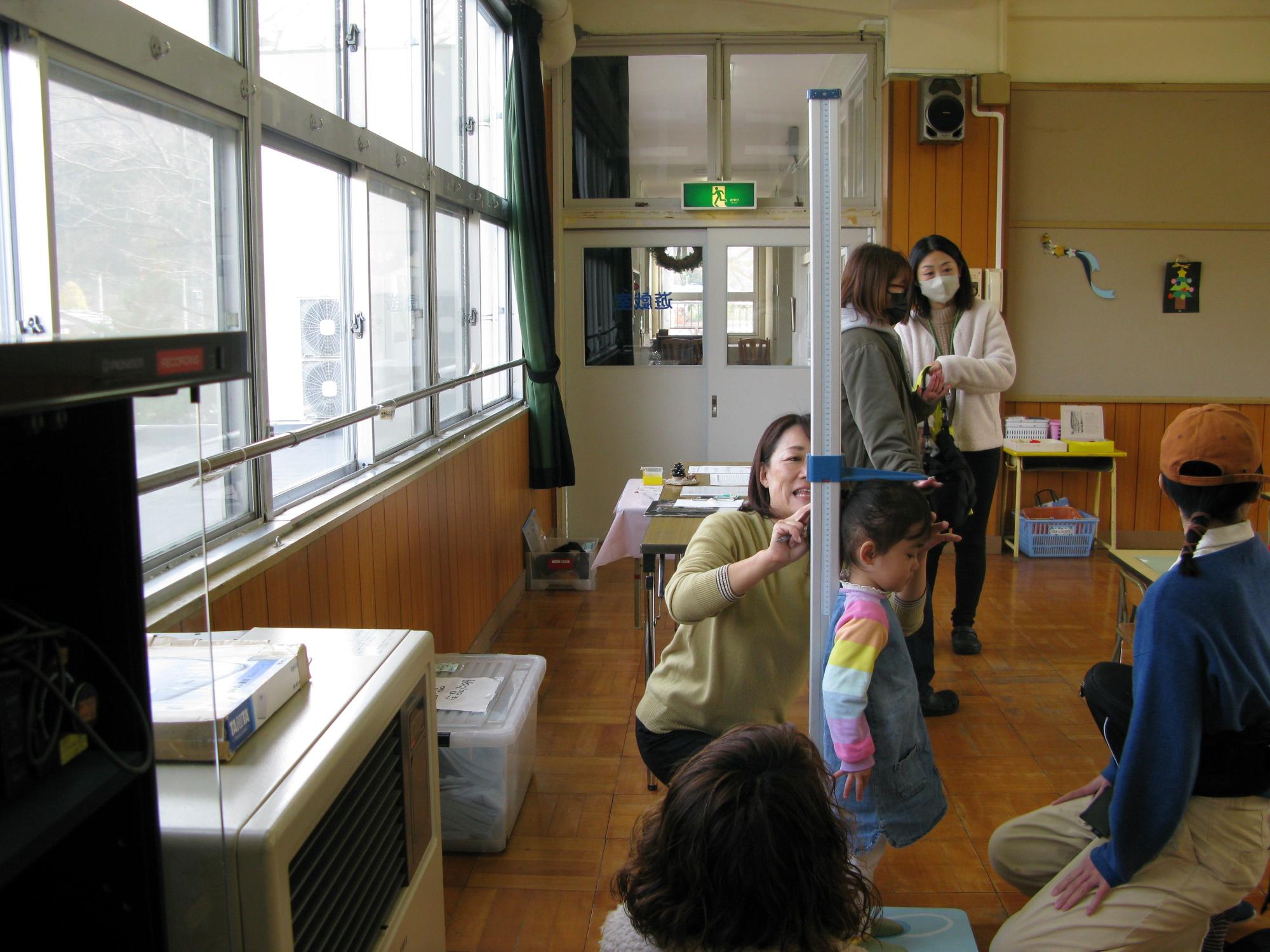 教室のような明るい部屋で、大人の女性たちが背の高い測定器を使って小さな女の子の身長を測っている様子の写真