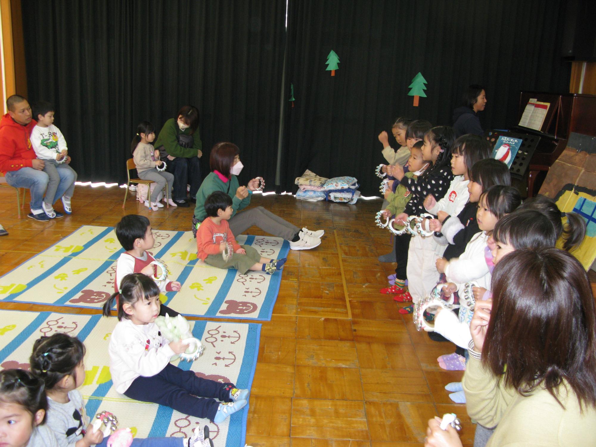 小さなクリスマスツリーの飾りがつけられた黒いカーテンが引かれた広い室内で、床に敷かれたマットの上で大勢の幼児たちが座り、向かい合って立つ園児たちが鈴を持って演奏を披露している様子の写真