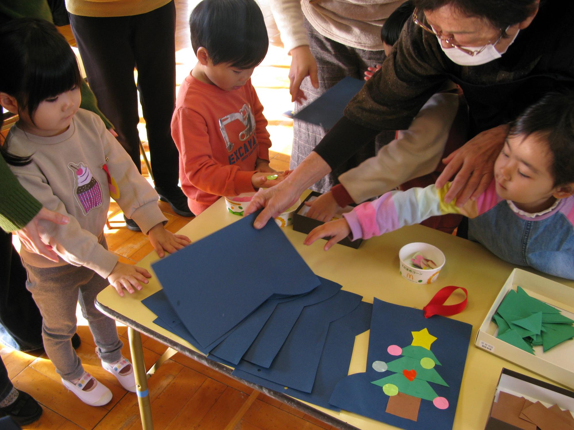 星と飾り付けがされた緑のクリスマスツリーの貼り絵をされた、青い画用紙が並んだ小さなテーブルを高齢の女性スタッフや保護者と思われる大人たちと一緒に数人の幼児たちが囲んでいる写真