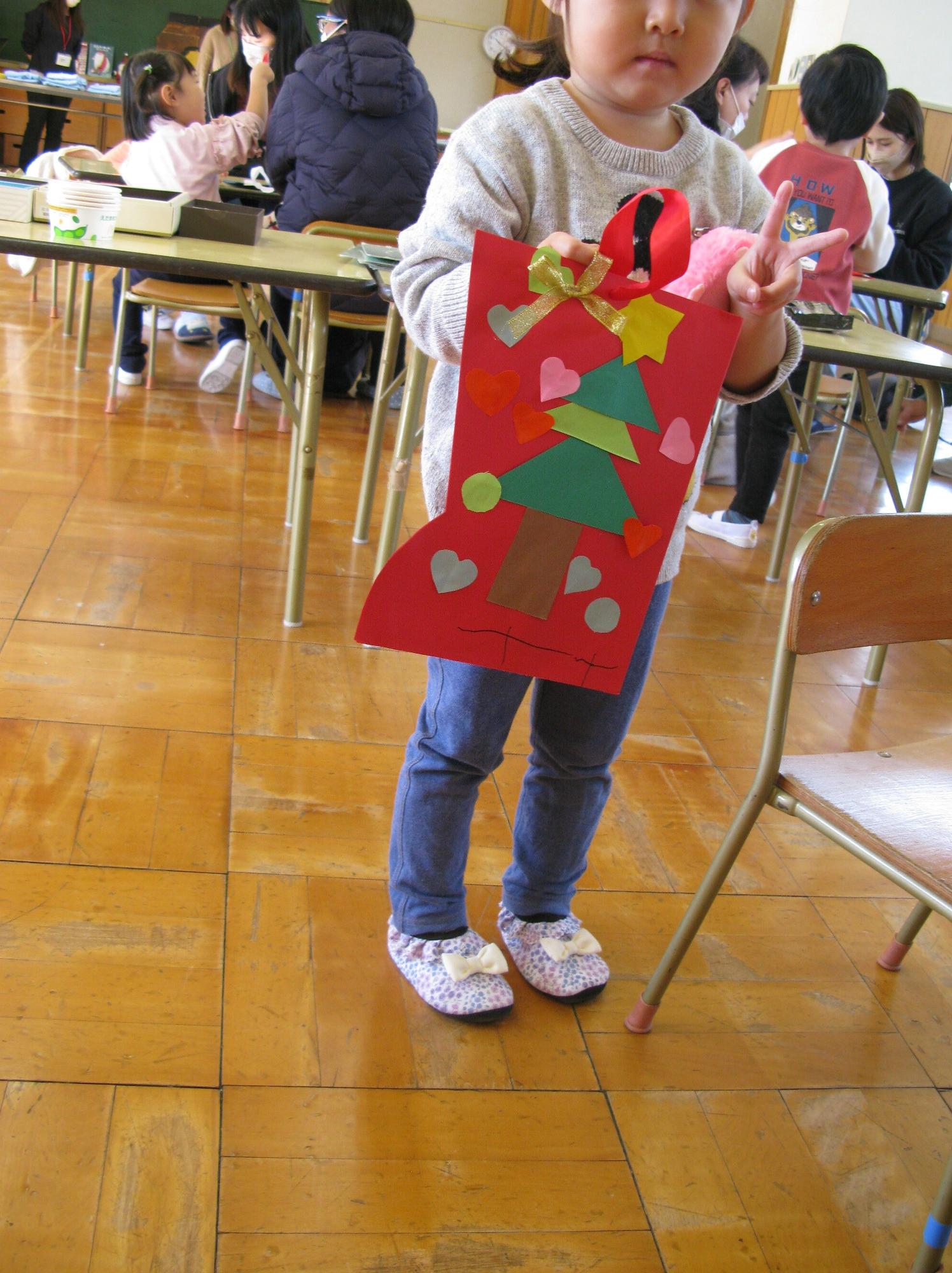室内で、赤い画用紙で作ったクリスマスブーツの飾りを、緑のツリーやハートのシールで飾り付けて完成させた作品を胸に抱えて立ち、ピースサインをしながらカメラに見せる幼い女の子の写真