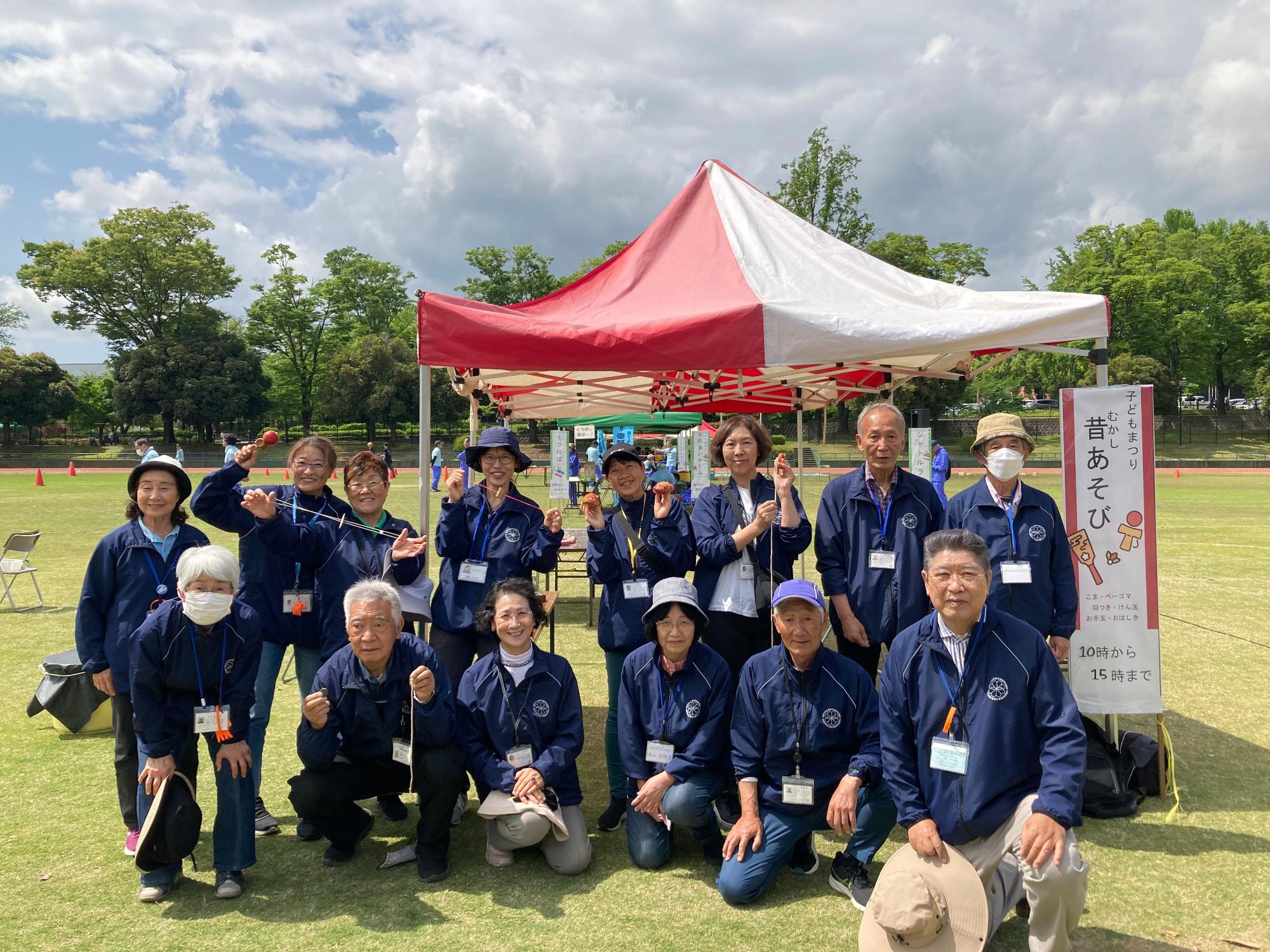 赤い帽子と青い上着を着用した男女が、屋外の広場で「昔あそび」と書かれた看板と赤いテントを背景に、楽しそうな表情で集合している写真