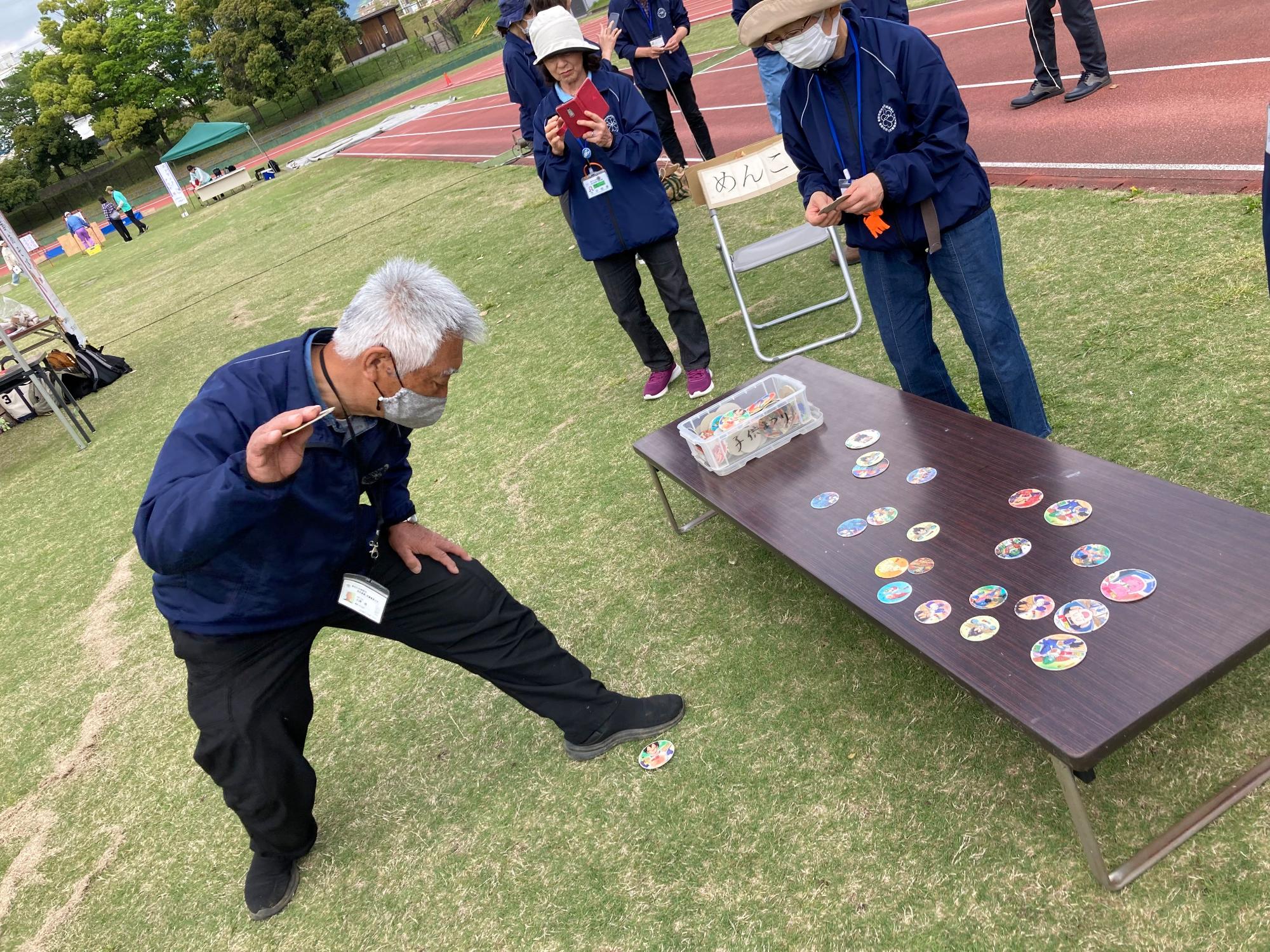屋外の陸上競技場で、紺色のジャンパーを着た男性が、しゃがんで「めんこ」で遊んでいる様子を捉えた写真